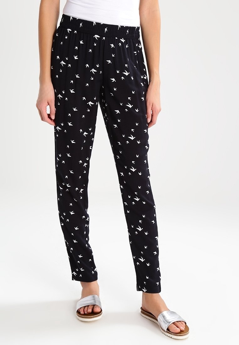 Černé kalhoty s bílými vzory ptáků, elastický pas, tapered střih, lehká látka, spárované se stříbrnými klouzacími sandály.