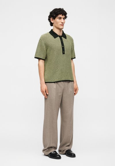 Άνδρας στέκεται φορώντας μια υφασμάτινη κοντομάνικη πράσινη και μαύρη πόλο μπλούζα, άνετο μπεζ παντελόνι και μαύρα παπούτσια πάνω σε λευκό φόντο.