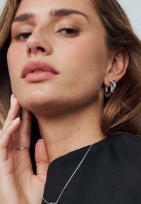 Silverfärgade hoopörhängen bärs, med en slät, polerad yta. Ett delikat silverfärgat kedjehalsband kompletterar en mörk tygblus.