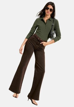 Grünes, geripptes Langarm-Poloshirt, braune, hochtaillierte Schlagjeans, brauner Gürtel, schwarze Handtasche und schwarze Sonnenbrille.