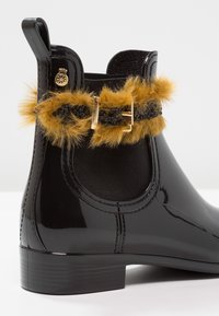 Botte de pluie noire avec une finition brillante, présentant un empiècement en fourrure beige, un panneau �élastique noir, et un accent en boucle dorée. Design à talon bas.