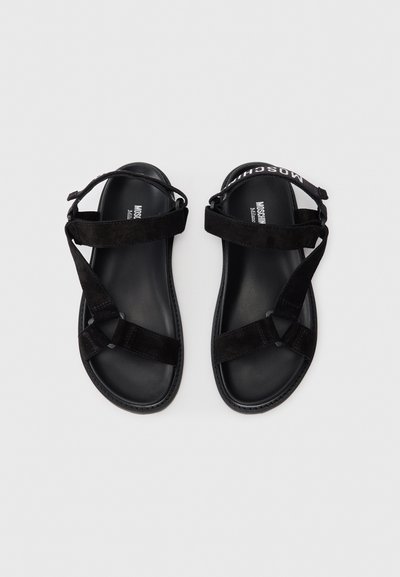 Sandale negre din suede cu talpă plată, cu bretele încrucișate și curea ajustabilă pentru gleznă. Logo pe curea în alb contrastant.