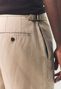 Beige Hose mit glatter Oberfläche, ausgestattet mit einer Gesäßtasche mit Knopf, Seitentaschen und einem verstellbaren Gürtelschlaufen-Detail.