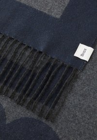 Coperte in lana blu scuro con motivo geometrico grigio, con bordi frangiati e un'etichetta nell'angolo. Texture morbida e aspetto caldo.