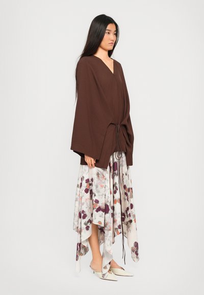 Woman with long dark hair wearing a brown loose top with tied front and a white floral asymmetrical skirt with pointed white heels.