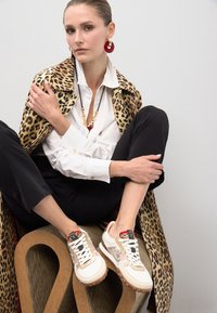 Zapatillas con parte superior de tela beige, acentos de estampado de leopardo, cordones blancos y suela de goma texturizada. El modelo se encuentra sobre un fondo de estampado de leopardo.