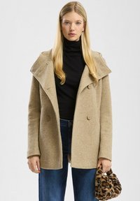 Beež tekstureeritud kahekordse nööbiga mantel suure kraega. Kantakse musta kõrge kaelusega särk üle, koos siniste teksade ja leopardimustriga käekotiga.