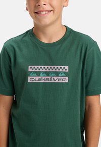 Zielona bawełniana koszulka z nadrukiem w wzór szachownicy oraz napisem „Quiksilver” w pastelowych różowych i niebieskich akcentach.