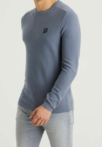 Man med en figurnära, ljusblå, strukturerad långärmad tröja med en liten svart logotyp på bröstet och ljusgrå jeans.