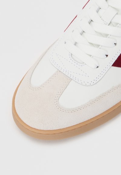 Valkoiset sneakerit, joissa on vaaleanruskea mokkanahkainen kärki, punainen yksityiskohta ja valkoiset nauhat. Ominaisuuksina sileä nahka ja teksturoitu mokkanahka kumipohjalla.