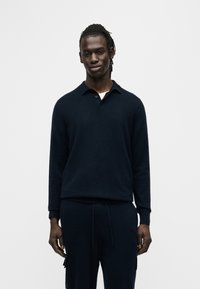 Πλεκτό πουλόβερ σε ναυτικό μπλε με γιακά και μακριά μανίκια, σε συνδυασμό με ταιριαστό ναυτικό μπλε παντελόνι jogger. Μαλακή υφή, μινιμαλιστικός σχεδιασμός.