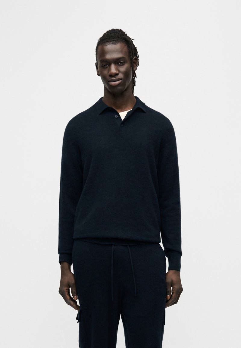 Πλεκτό πουλόβερ σε ναυτικό μπλε με γιακά και μακριά μανίκια, σε συνδυασμό με ταιριαστό ναυτικό μπλε παντελόνι jogger. Μαλακή υφή, μινιμαλιστικός σχεδιασμός.