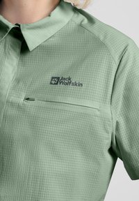 Camicia a maniche corte verde chiaro con motivo a griglia, dotata di tasca frontale e logo sul petto sinistro. Tessuto liscio.