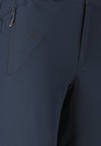 Gros plan sur un pantalon d'extérieur bleu foncé montrant une poche zippée à l'avant et un petit logo noir près de la hanche.