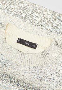 Suéter plateado con lentejuelas, con un cuello de crema texturizado y acanalado, y una etiqueta de cuidado visible en el collar. Superficie lisa con toques brillantes.