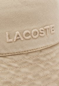 Beige cap made from textured fabric featuring embroidered "LACOSTE" logo in light beige thread, with a structured design and curved brim.