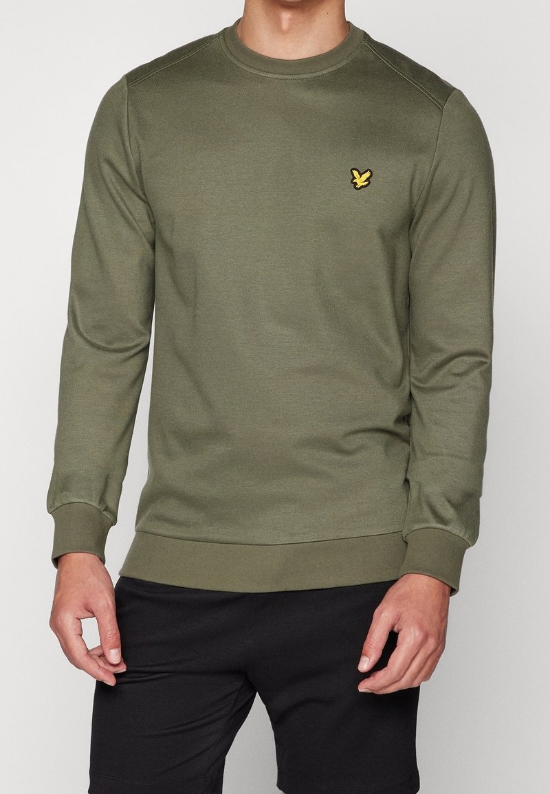 Homme portant un sweat-shirt à manches longues vert olive avec un logo d'oiseau jaune sur la poitrine et un short noir, debout devant un fond blanc.