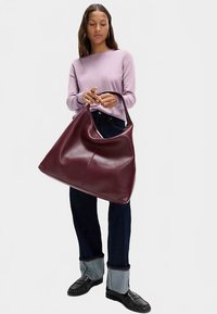 Stor vinröd läderväska med en mjuk, slät yta och en böjd silhuett. Väskan har ett enda övre handtag och bärs av en person.