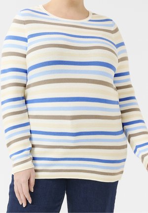 Kvinde iført en langærmet striktrøje med vandrette beige, blå, lyseblå og brune striber, kombineret med mørkeblå bukser.