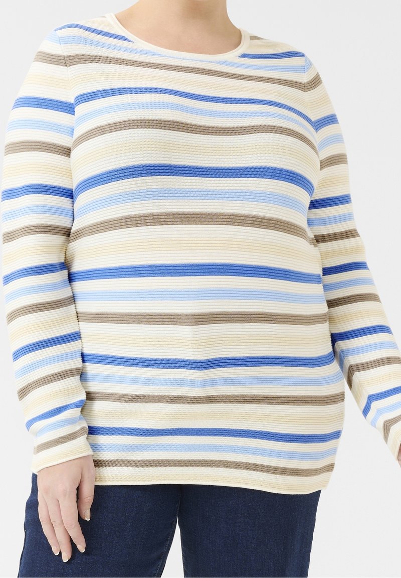 Femme portant un pull en maille à manches longues avec des rayures horizontales beige, bleu, bleu clair et marron, associé à un pantalon bleu foncé.