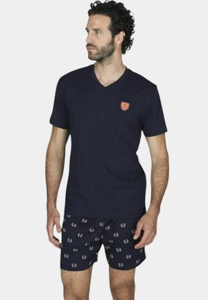 Uomo con capelli ricci e scuri che indossa una T-shirt blu navy con scollo a V e pantaloncini blu navy con un motivo ripetuto di granchi chiari.