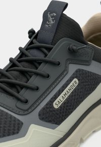 Γκρι και μαύρο αθλητικό παπούτσι με πλέγμα και συνθετικά υλικά, υφές, στενές κορδόνες και έντονη ετικέτα λογότυπου.