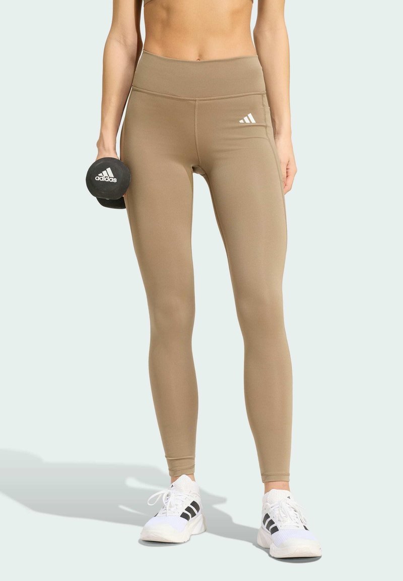 Vrouw draagt beige Adidas legging en witte Adidas sneakers, houdt een zwarte Adidas dumbbell in haar linkerhand.