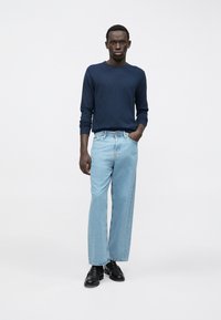 Νεαρός άνδρας στέκεται με το ένα χέρι στην τσέπη, φορώντας ναυτικό πουλόβερ, ανοιχτό μπλε φαρδιά τζιν και μαύρα παπούτσια μπροστά σε απλή φόντο.