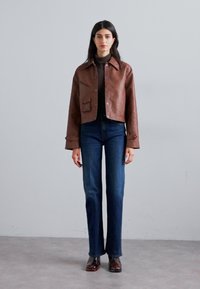 Chaqueta recortada de cuero marrón con cuello puntiagudo, bolsillo frontal y detalle de botones, combinada con unos pantalones vaqueros acampanados de color azul oscuro y zapatos brillantes.
