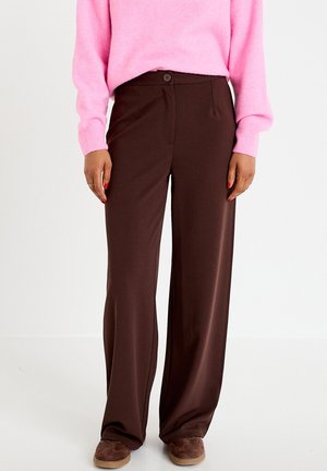 Personne portant un pantalon large marron foncé, un pull rose à manches longues et des chaussures marron, debout devant un fond uni.