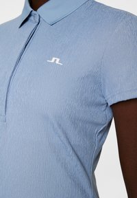 Jasnoniebieska, teksturowana koszulka polo z kołnierzykiem, z białym logo na klatce piersiowej i krótkimi rękawami.