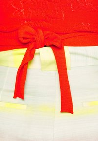 Haut rouge en maille noué en nœud à la taille sur un pantalon blanc et jaune pâle rayé.