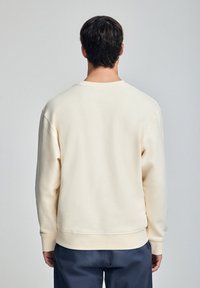 Felpa crema con scollo a girocollo, polsini e orlo a costine, e texture morbida, vista da dietro. Indossata con pantaloni larghi scuri.