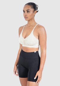 Beige urheiluliivi, jossa on rypytetty etuosa, yhdistettynä korkeavyötäröisiin mustiin shortseihin. Molemmat on valmistettu joustavasta, hengittävästä kankaasta.