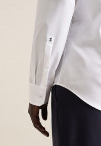 Camicia bianca a maniche lunghe con polsini a bottone, caratterizzata da un piccolo ricamo floreale nero sulla manica. Tessuto morbido, vestibilità tailored.