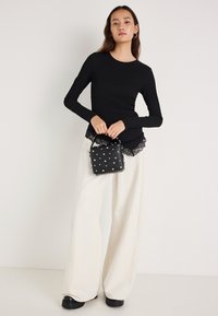 Schwarzes, geripptes Langarmshirt mit Spitzenabschluss, kombiniert mit weißen, weit geschnittenen Hosen. Hält eine kleine schwarze Handtasche mit silbernen Verzierungen.