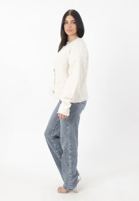 Maglione di colore crema, con texture, maniche lunghe e tasche frontali, abbinato a jeans dritti blu chiaro e sandali con tacco trasparente.