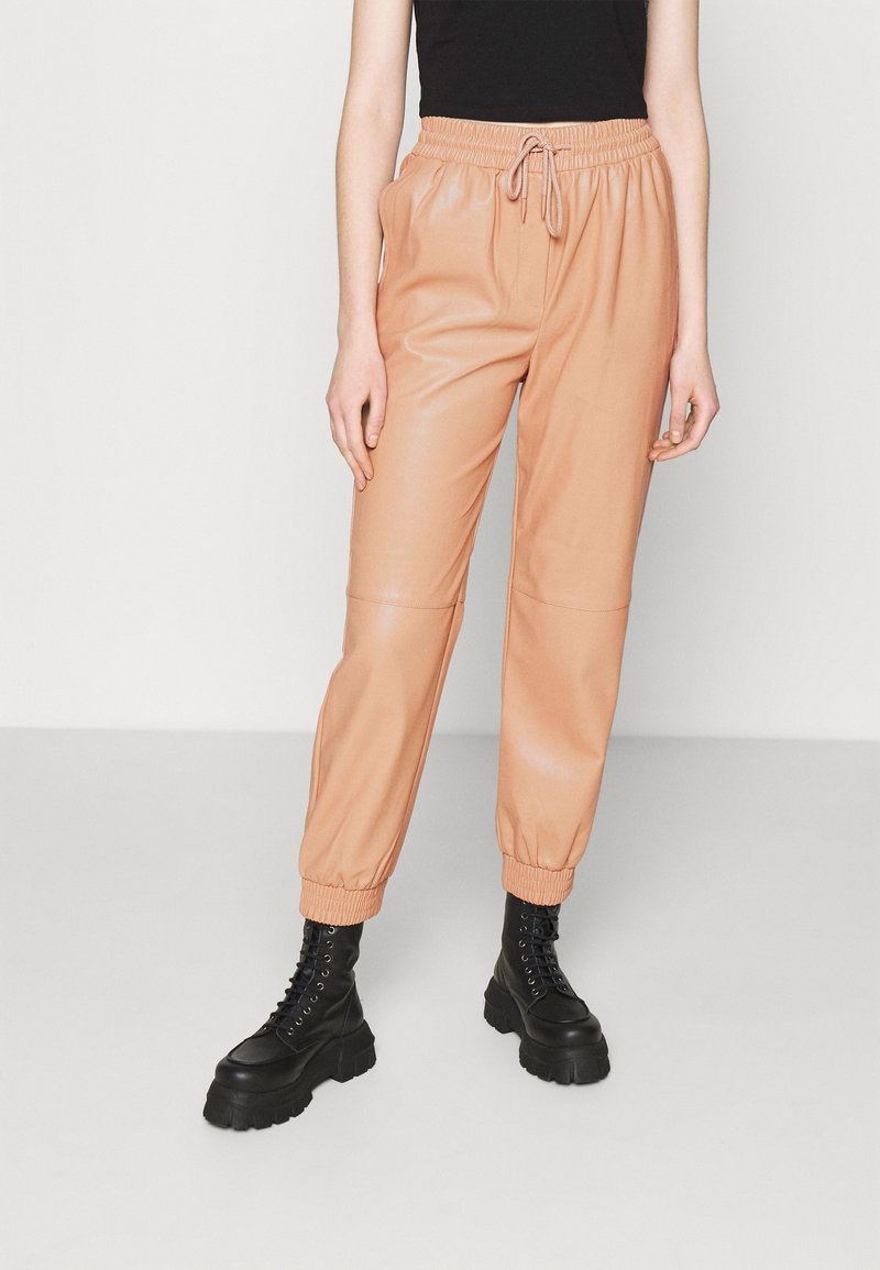 Hellbeige Jogginghose aus Kunstleder mit elastischem Taillenbund und Bündchen, ausgestattet mit einem Kordelzug und Nahtdetails. Kombiniert mit schwarzen Stiefeln im Kampfstil.