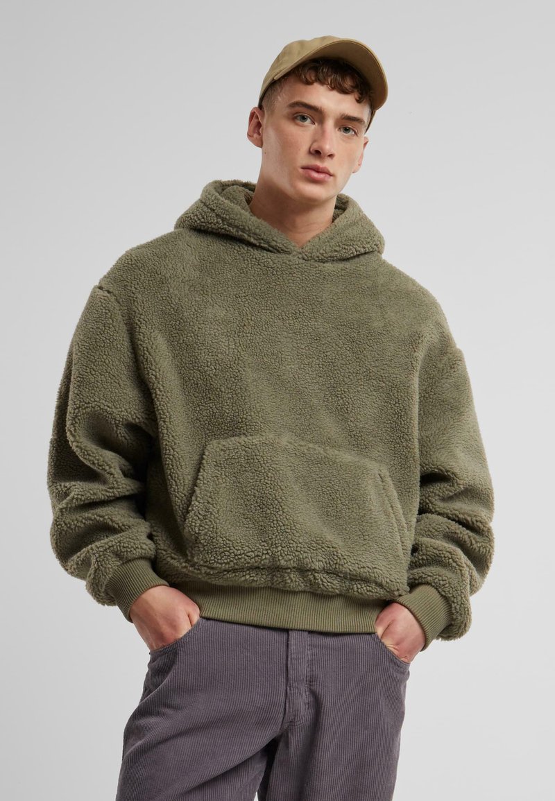 Olivenfarbener Fleece-Hoodie mit Kängurutasche, strukturiertem Stoff, gerippten Bündchen und Saum, kombiniert mit grauen Cordhosen.