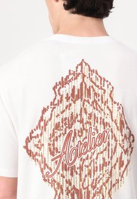 Camiseta blanca de algodón con un estampado decorativo texturizado en rojo y dorado, que muestra la palabra "Atelier" en una fuente estilizada.