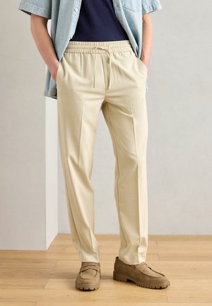 Beige skräddarsydda byxor med elastisk midja och dragsko. Kombinerade med en marinblå topp och en ljus denimskjorta; bärs med bruna loafers.