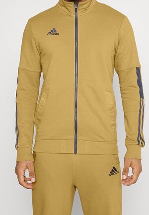 Mężczyzna w musztardowo-żółtym dresie Adidas z kurtką na suwak i pasującymi spodniami z czarnymi logo Adidas i czarnymi paskami na rękawach.