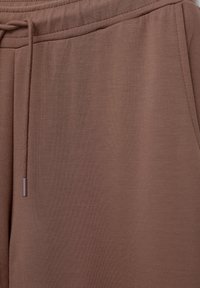 Pantaloni de trening din bumbac maro, cu un corset elastic și un șnur care atârnă pe partea stângă.