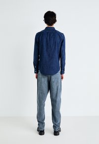 Camisa de denim en azul oscuro con mangas largas, ajuste ajustado y puños con botón, combinada con jeans ligeros de corte holgado en azul y zapatillas negras.
