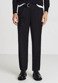 Mann trägt schwarze gerade geschnittene Hose mit einem schwarzen Gürtel und schwarzen Schnürschuhen vor einem einfarbigen Hintergrund.