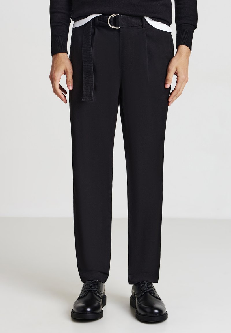 Mann trägt schwarze gerade geschnittene Hose mit einem schwarzen Gürtel und schwarzen Schnürschuhen vor einem einfarbigen Hintergrund.