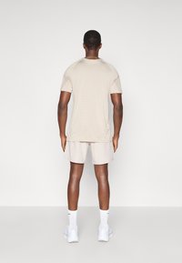 Άνδρας που στέκεται με την πλάτη γυρισμένη, φορώντας αθλητικό μπλουζάκι κοντομάνικο σε ανοιχτό μπεζ χρώμα, αντίστοιχες βερμούδες, λευκές κάλτσες και λευκά αθλητικά παπούτσια, σε απλό φόντο.