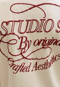 Broderie rouge en cursive et à bloc indiquant « STUDIO By original Crafted Aesthetics » sur un tissu crème avec des boucles décoratives.