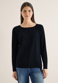 Femme aux cheveux longs et foncés, portant un pull noir à manches longues et un jean bleu, debout devant un fond uni.