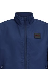 Marineblaue Jacke aus glattem Stoff, mit hohem Kragen und Reißverschluss vorne, versehen mit einem schwarzen Etikett mit dem Text "RETOUR."